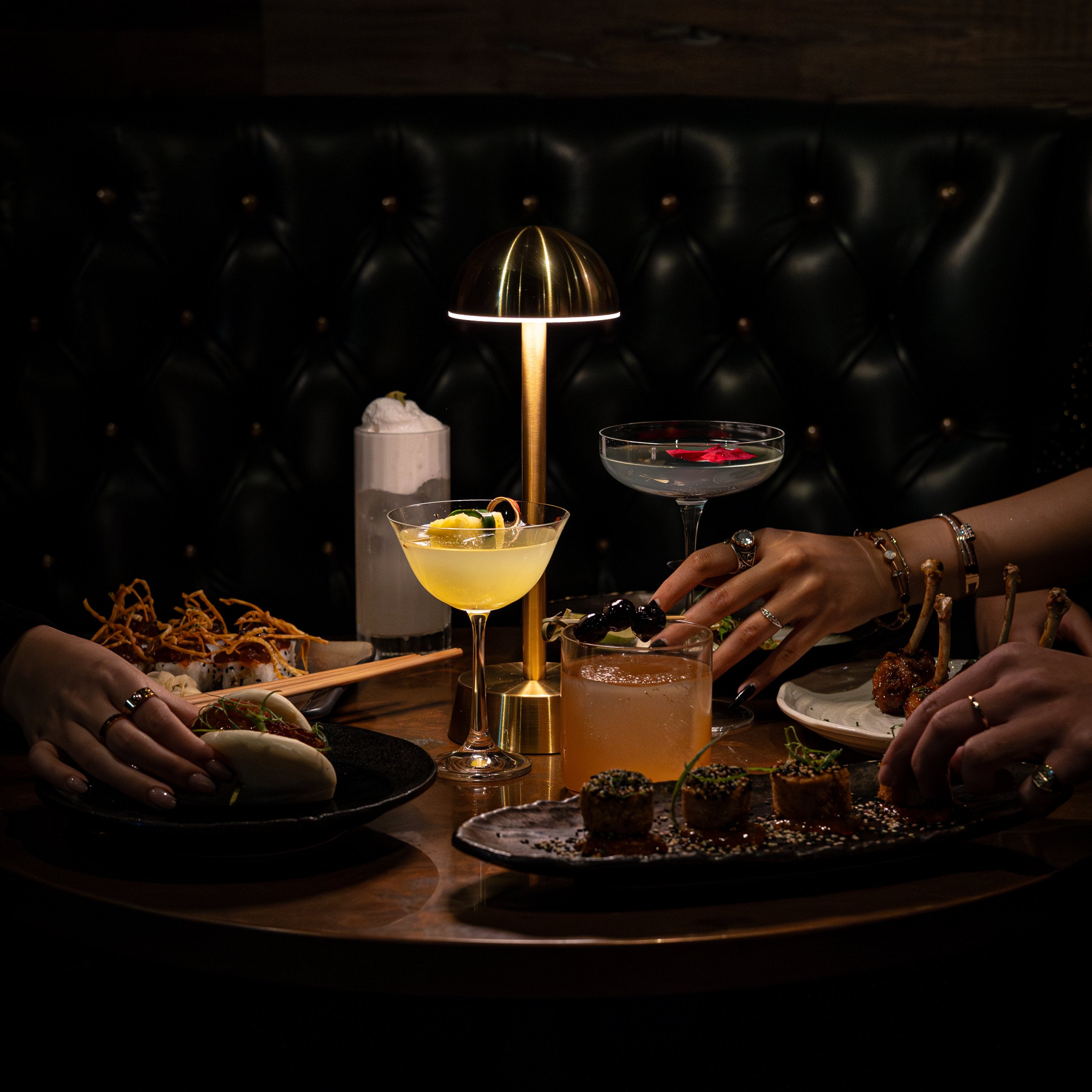 A bar table with exciting food and cocktails illuminated by a brass cordless table lamp.