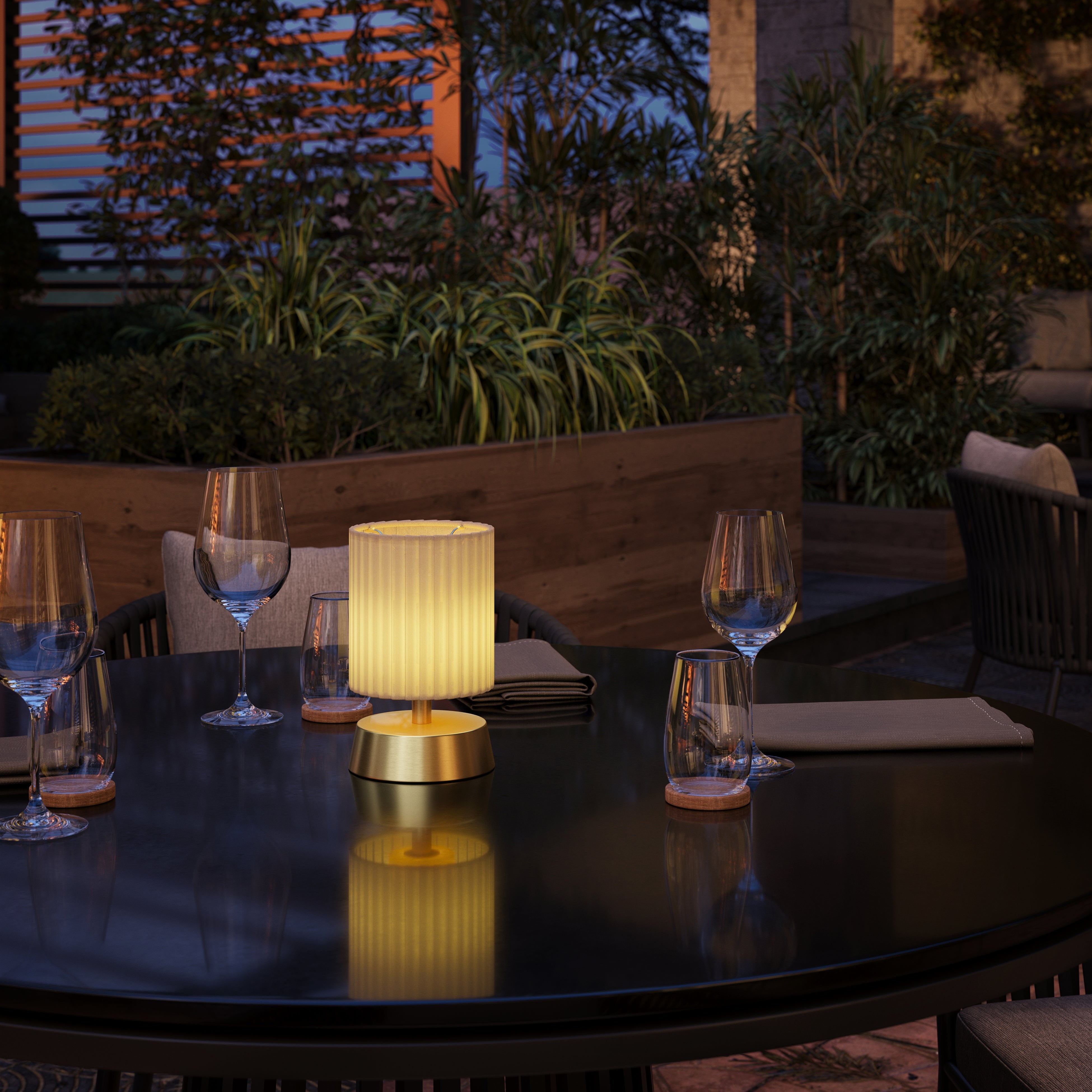 A small rechargeable lamp with a brass base finished with an ivory pleated shade on an outdoor dining table.
