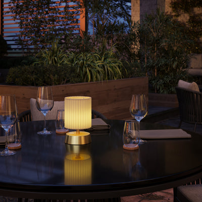A small rechargeable lamp with a brass base finished with an ivory pleated shade on an outdoor dining table.
