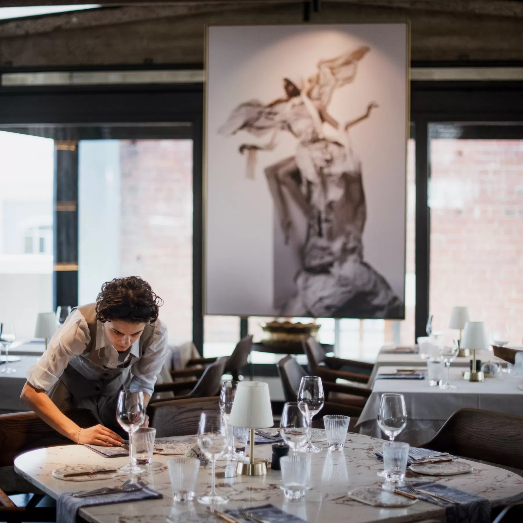 Person setting a table in a restaurant with cordless table lamps and a large abstract painting on the wall.
