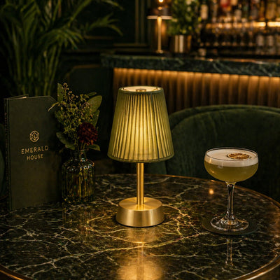 Gold rechargeable lamp on a marble table with a cocktail and 'Emerald House' menu in a bar setting