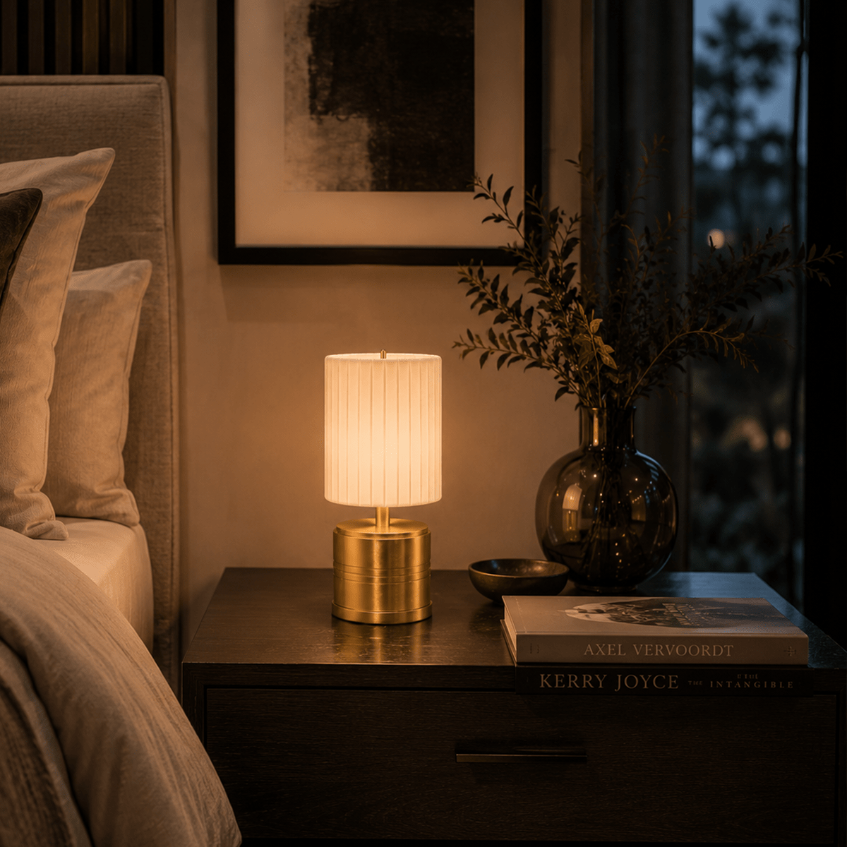 Rechargeable lamp on a bedside table with a book and plant in a dimly lit room.