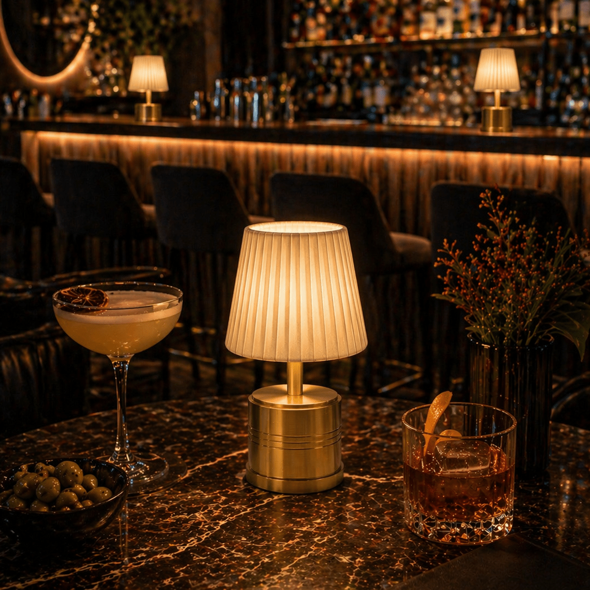 Cocktail glasses on a bar counter with a rechargeable cordless lamp and decorative elements in a dimly lit bar setting.