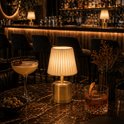 Cocktail glasses on a bar counter with a rechargeable cordless lamp and decorative elements in a dimly lit bar setting.