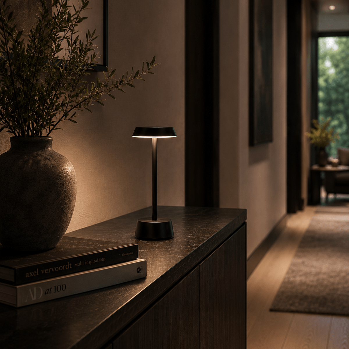 Modern rechargeable table lamp on a wooden surface with books and a vase in a dimly lit room.