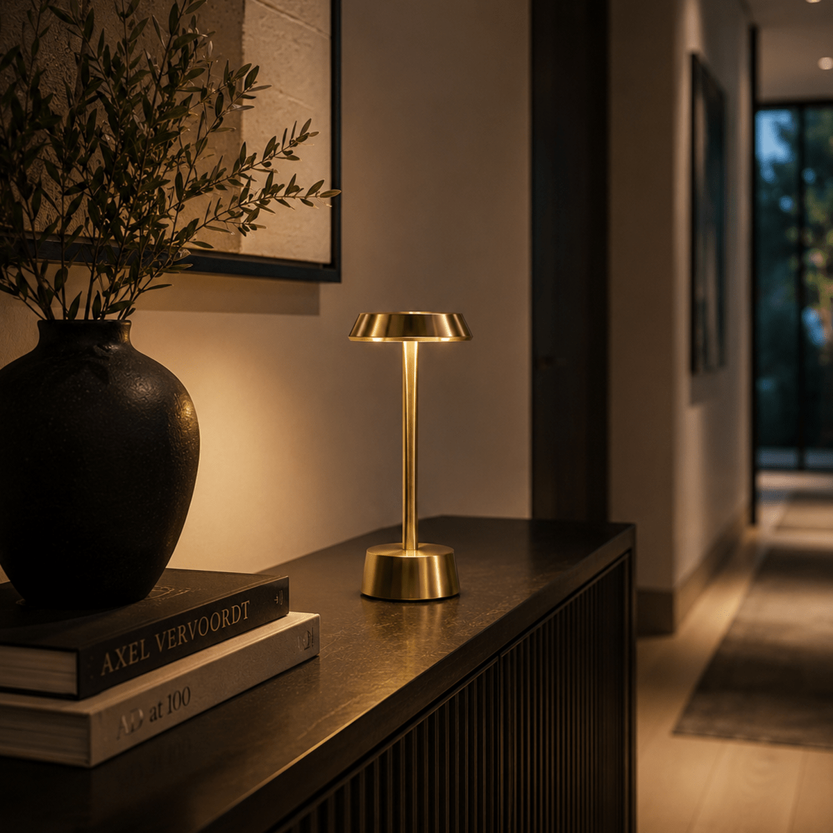 Gold rechargeable table lamp on a wooden surface with books and a vase in a dimly lit room.