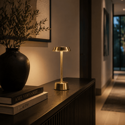 Gold rechargeable table lamp on a wooden surface with books and a vase in a dimly lit room.