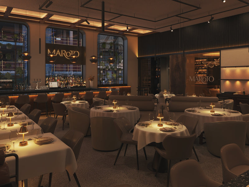 A fine dining restaurant with cordless table lamps, wine glasses and plates set for evening service. A bar in the background with ambient lighting around.